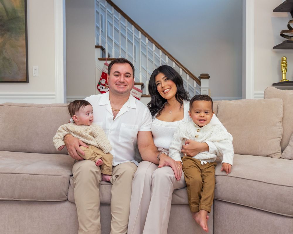 A family of four sits together on a beige couch in their living room with a staircase visible in the background.