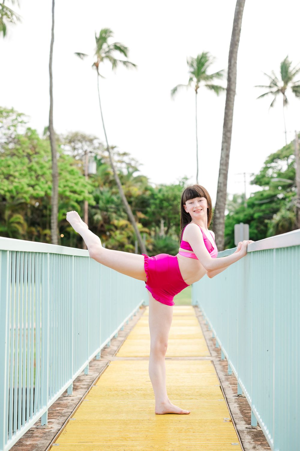 A dancer in pink performs a graceful pose on a tropical bridge pathway.