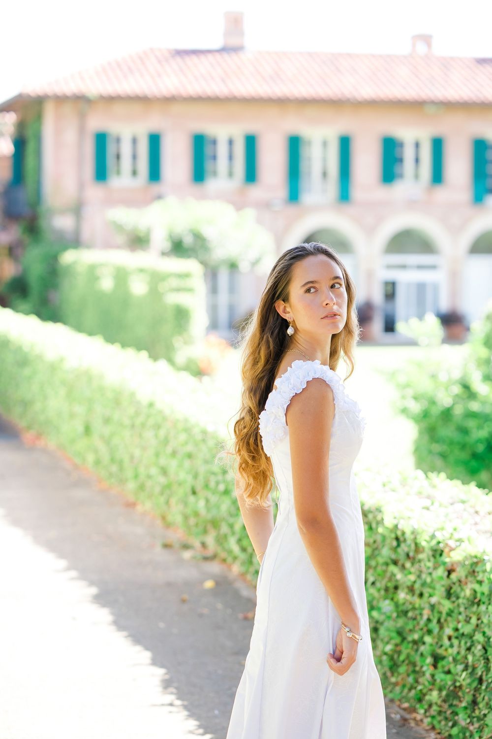 A person in an elegant white dress poses in front of a Mediterranean-style villa with blue shutters and manicured hedges.