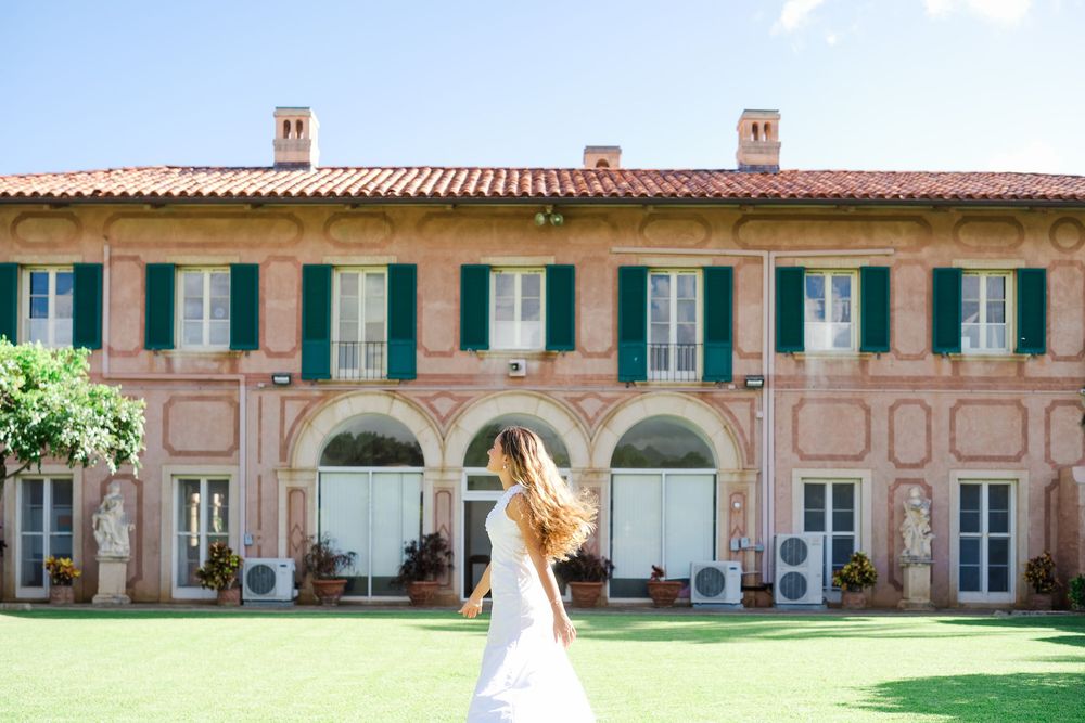 Person in white dress twirls on green lawn in front of pink Italian villa with green shuttered windows.
