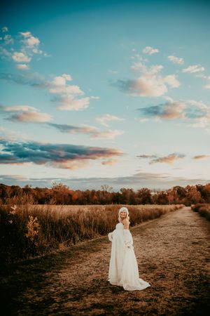 A person in a white dress stands on a dirt path at sunset with golden autumn fields and blue sky in the background.