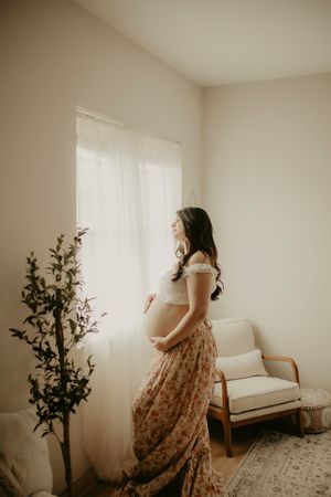 Pregnant woman in floral skirt and white lace top cradling her baby bump by a bright window in a serene room.