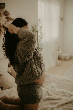 Person wearing cozy knit sweater and shorts relaxes on bed in warm sunlit room with minimalist decor.
