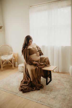 Indoor maternity session with soft natural light and flowing brown dress.