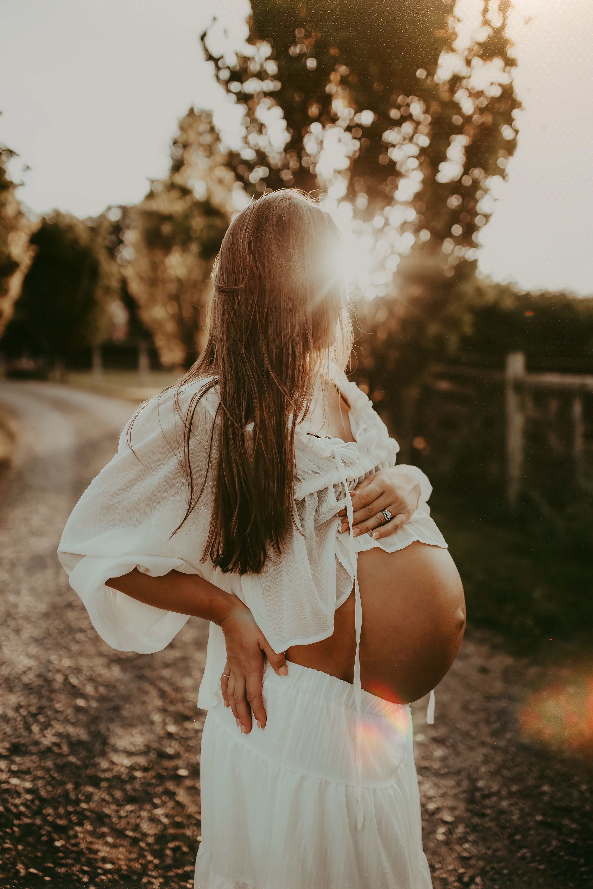 Pregnant woman in white crop top and skirt cradling her baby bump outdoors at golden sunset hour.