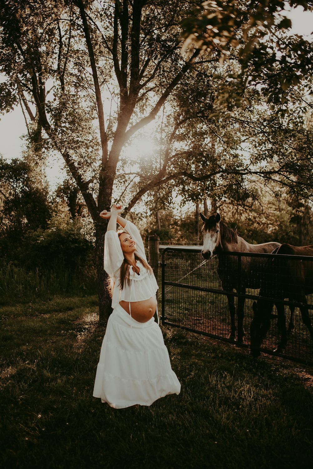 Pregnant woman in white dress poses gracefully outdoors at golden hour near a horse and metal fence.
