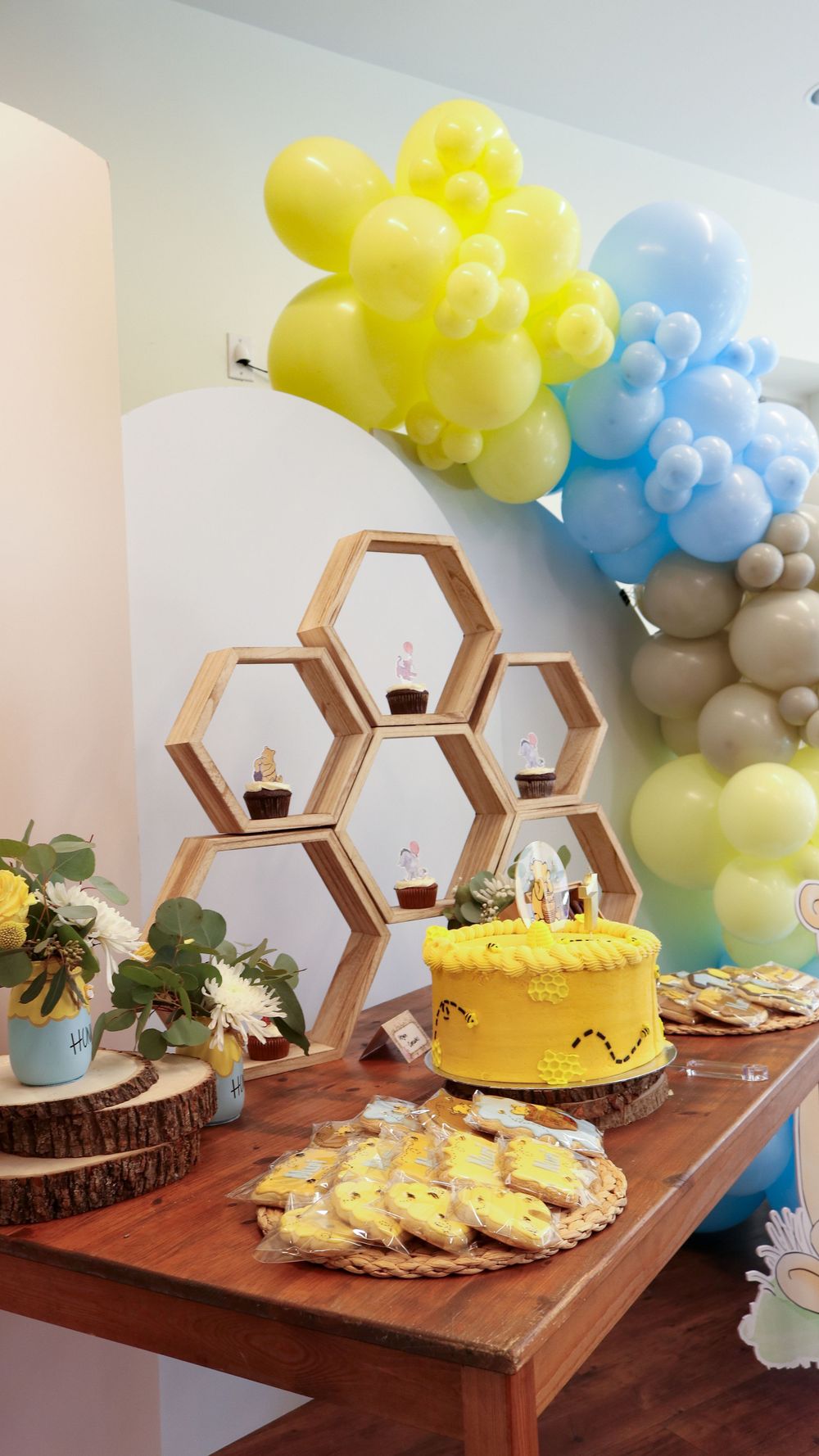 Bee-themed baby shower dessert table with yellow cake, honeycomb shelves, cookies, and colorful balloon arch.