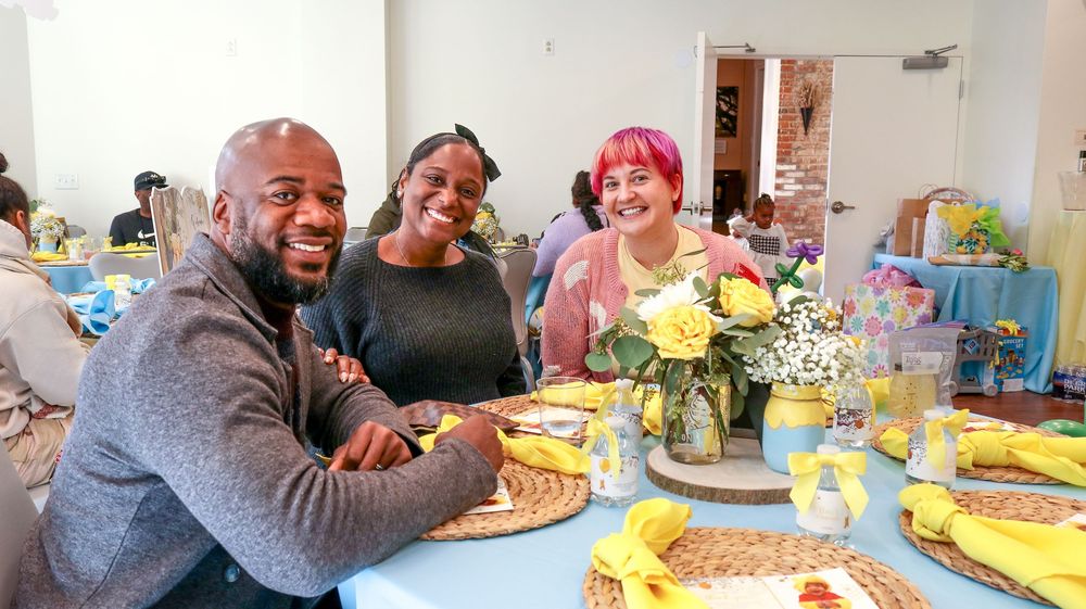 Three smiling guests sit together at a decorated table with yellow flowers at a festive baby shower event.