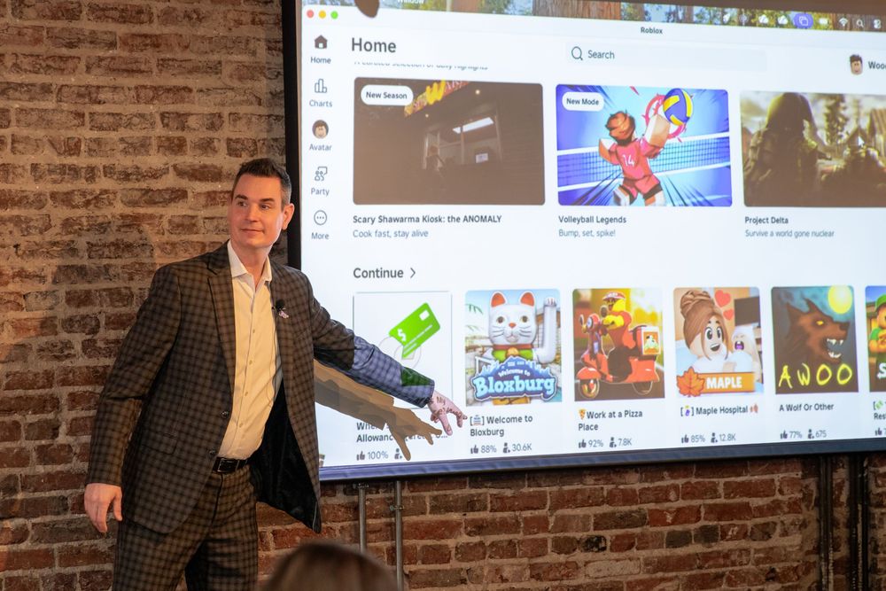 Professional speaker presenting to an audience in front of digital screens showing marketing content in a brick-walled venue.
