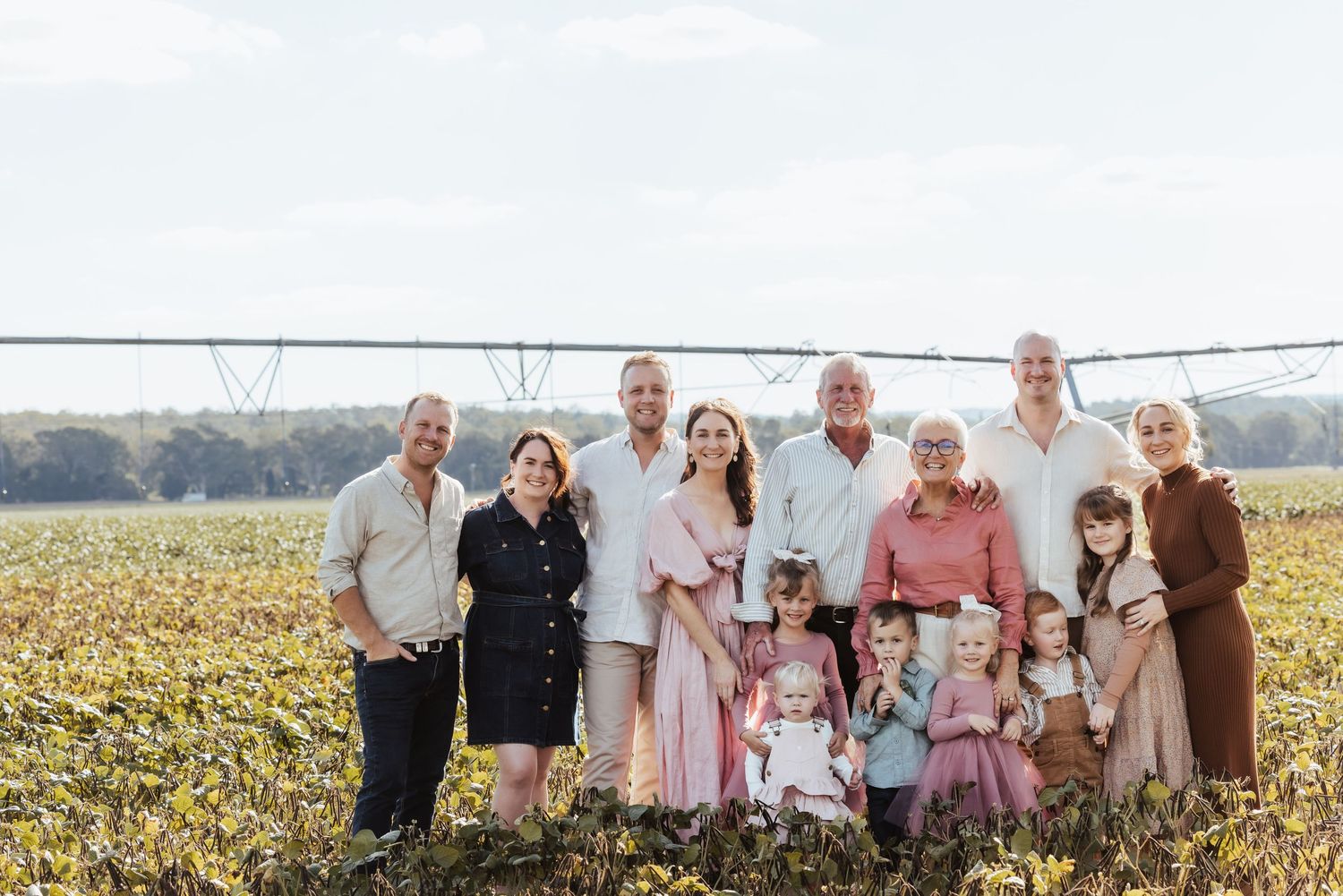 Large multigenerational family posing together in a sunny farm field with irrigation equipment in the background.