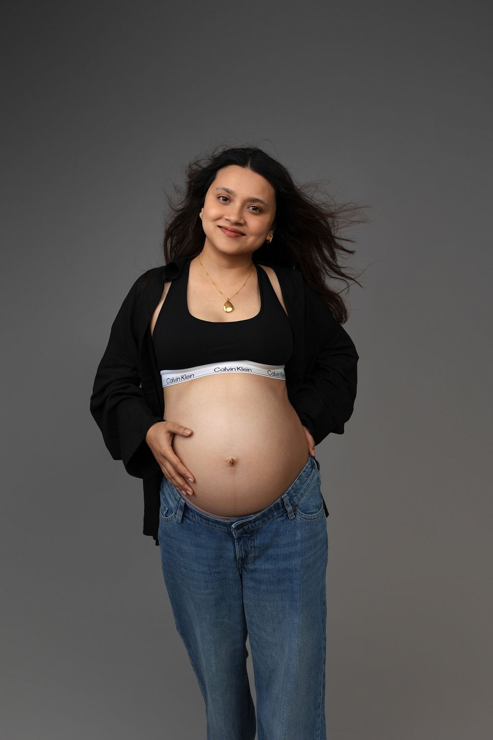 Smiling pregnant woman in Calvin Klein sports bra, black jacket, and jeans cradling her baby bump against gray background.