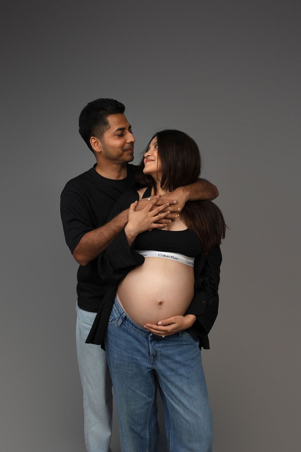 Couple embracing during maternity photo shoot, pregnant woman in Calvin Klein bra and jeans with partner in black outfit.