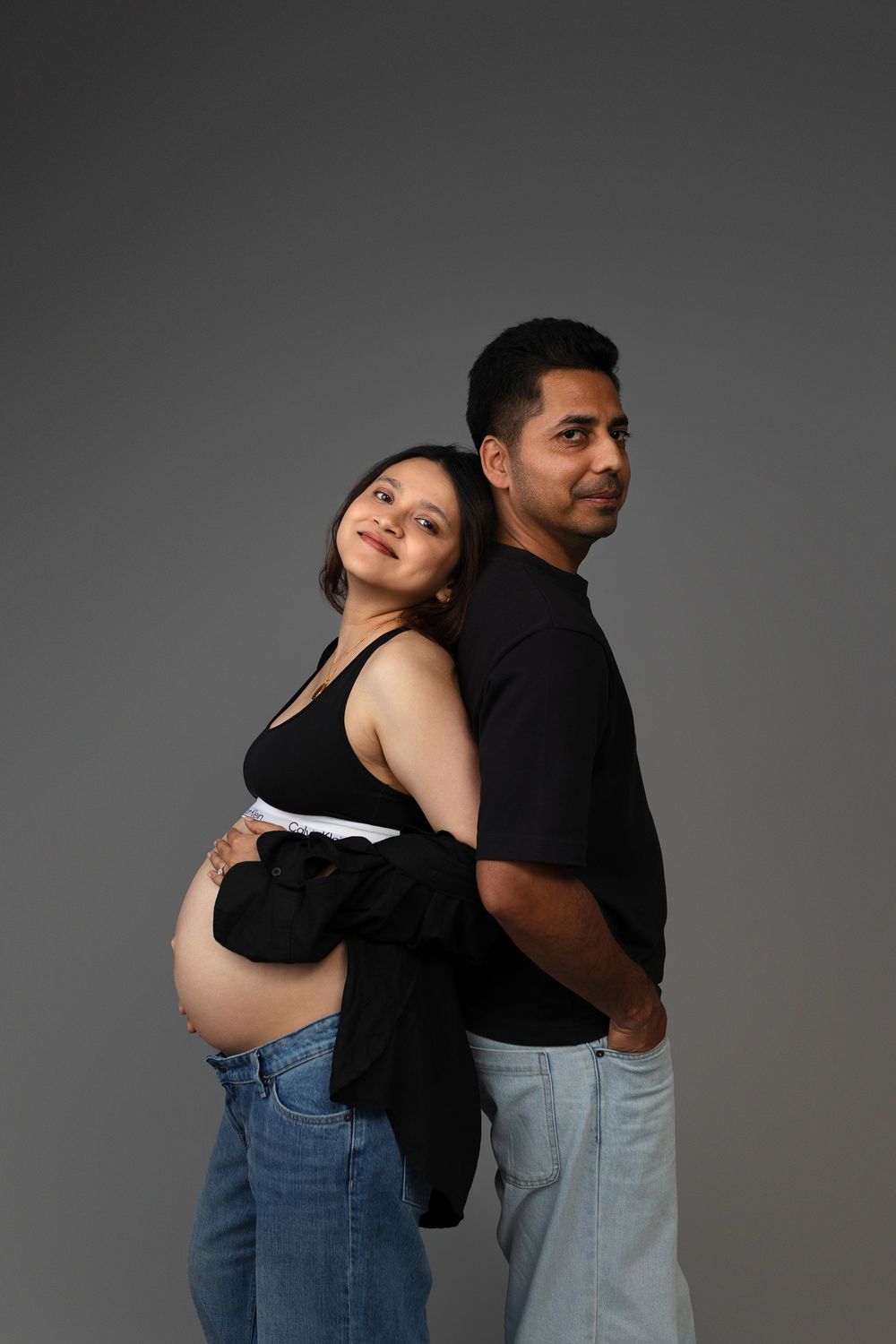 Pregnant woman in black top and jeans stands back-to-back with man in black shirt against gray background.