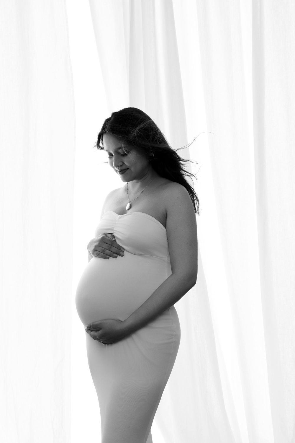 Black and white portrait of a pregnant woman in a strapless dress cradling her baby bump against white curtains.
