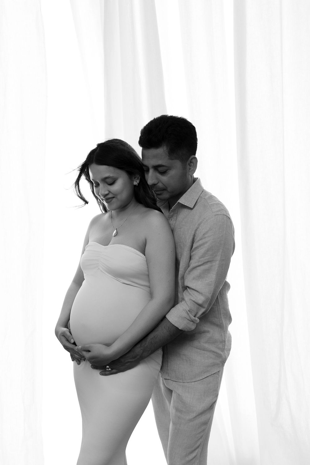 Black and white maternity photo of a couple, man embracing pregnant woman from behind, both looking down tenderly.