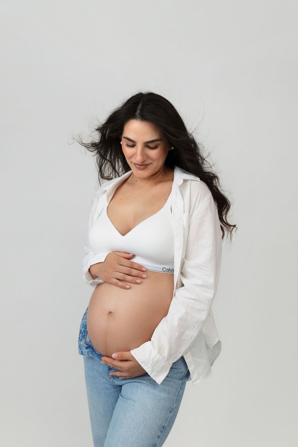 Smiling pregnant woman in white crop top and jeans cradling her baby bump against a light gray background.