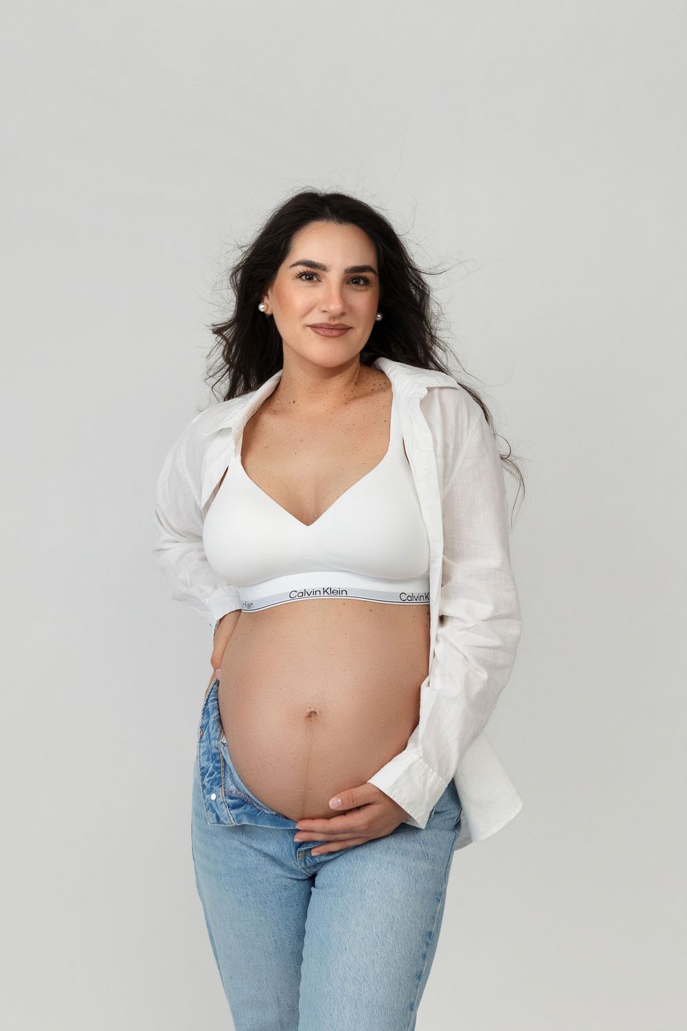 Pregnant woman in Calvin Klein bralette, open white shirt, and jeans cradling her baby bump against a light background.