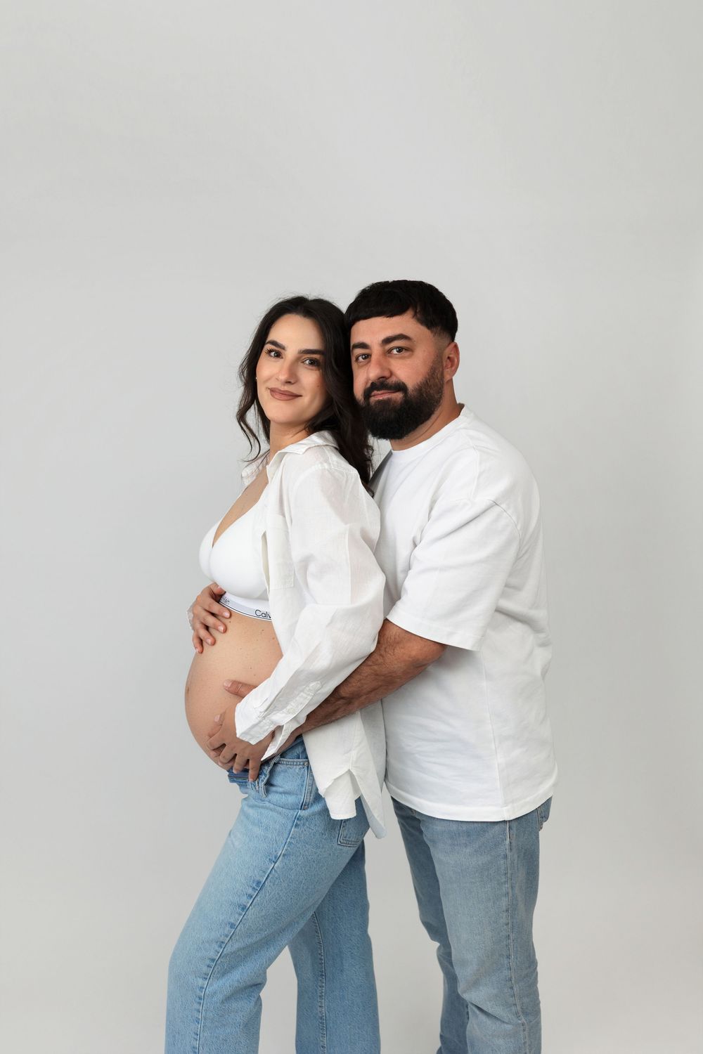 Pregnant couple in white shirts and jeans posing together, man embracing woman from behind on gray background.
