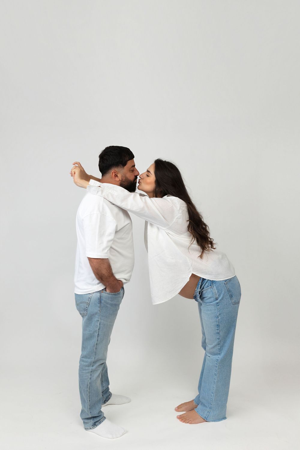 Couple in matching white shirts and jeans sharing a romantic kiss against a light gray background.