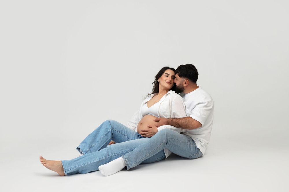 Couple sitting on white floor, pregnant woman leaning on man, both wearing jeans and white tops, maternity photo.