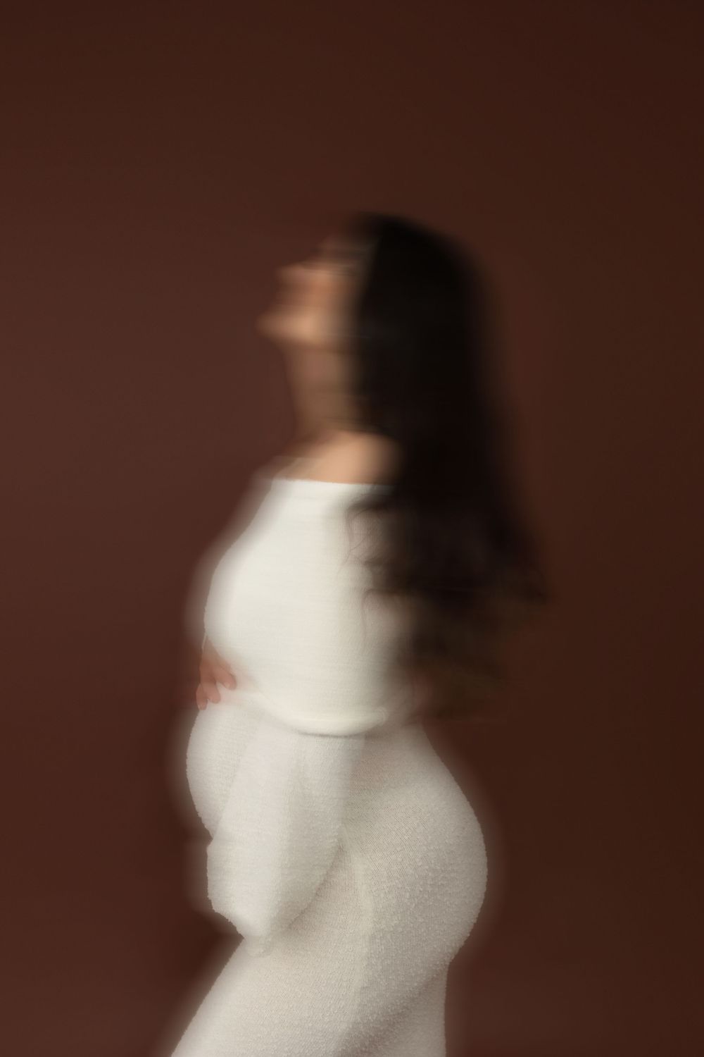 Blurred artistic portrait of a pregnant woman in white dress cradling her belly against a brown background.