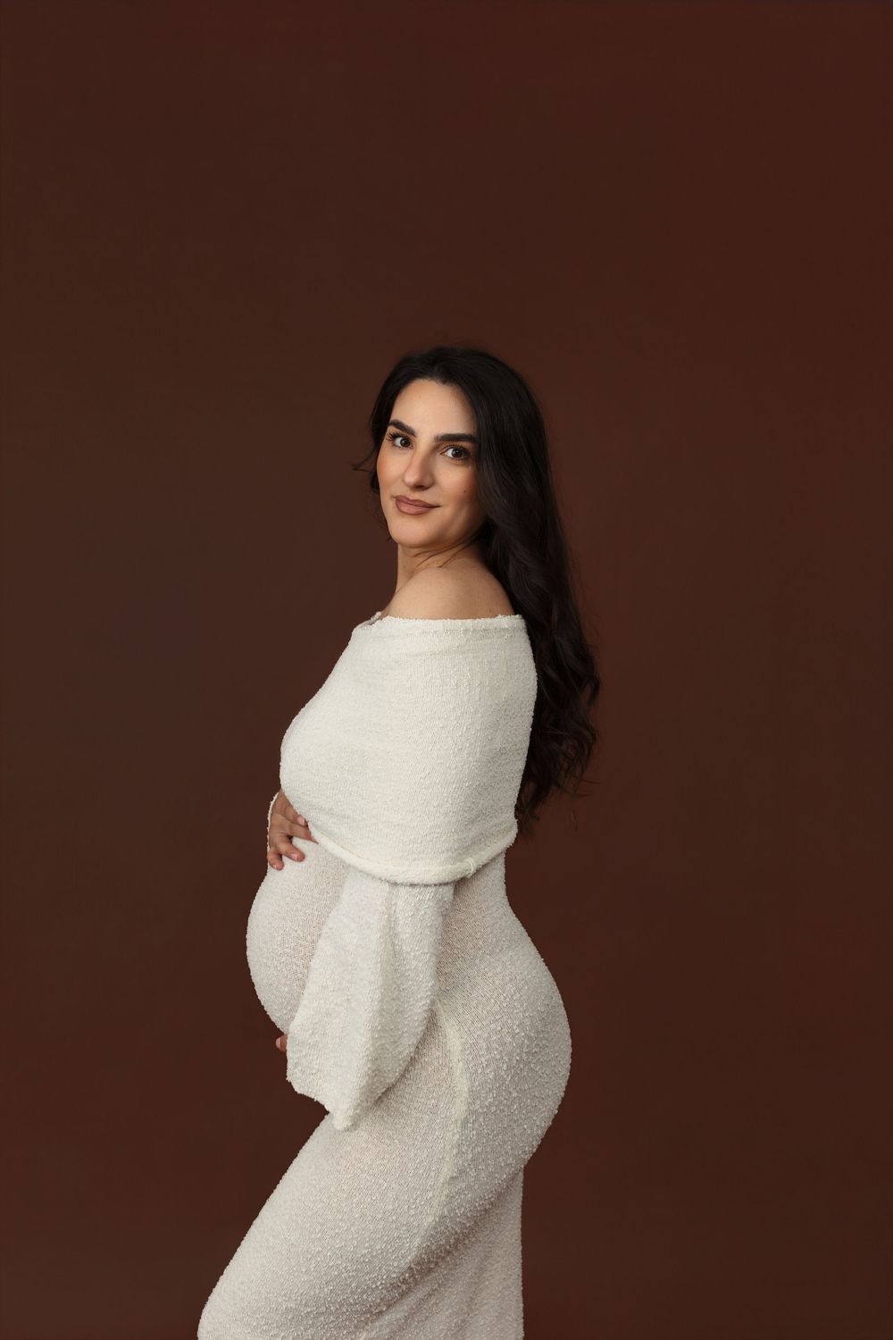 Pregnant woman in white off-shoulder dress posing sideways on brown background, cradling baby bump with gentle smile.