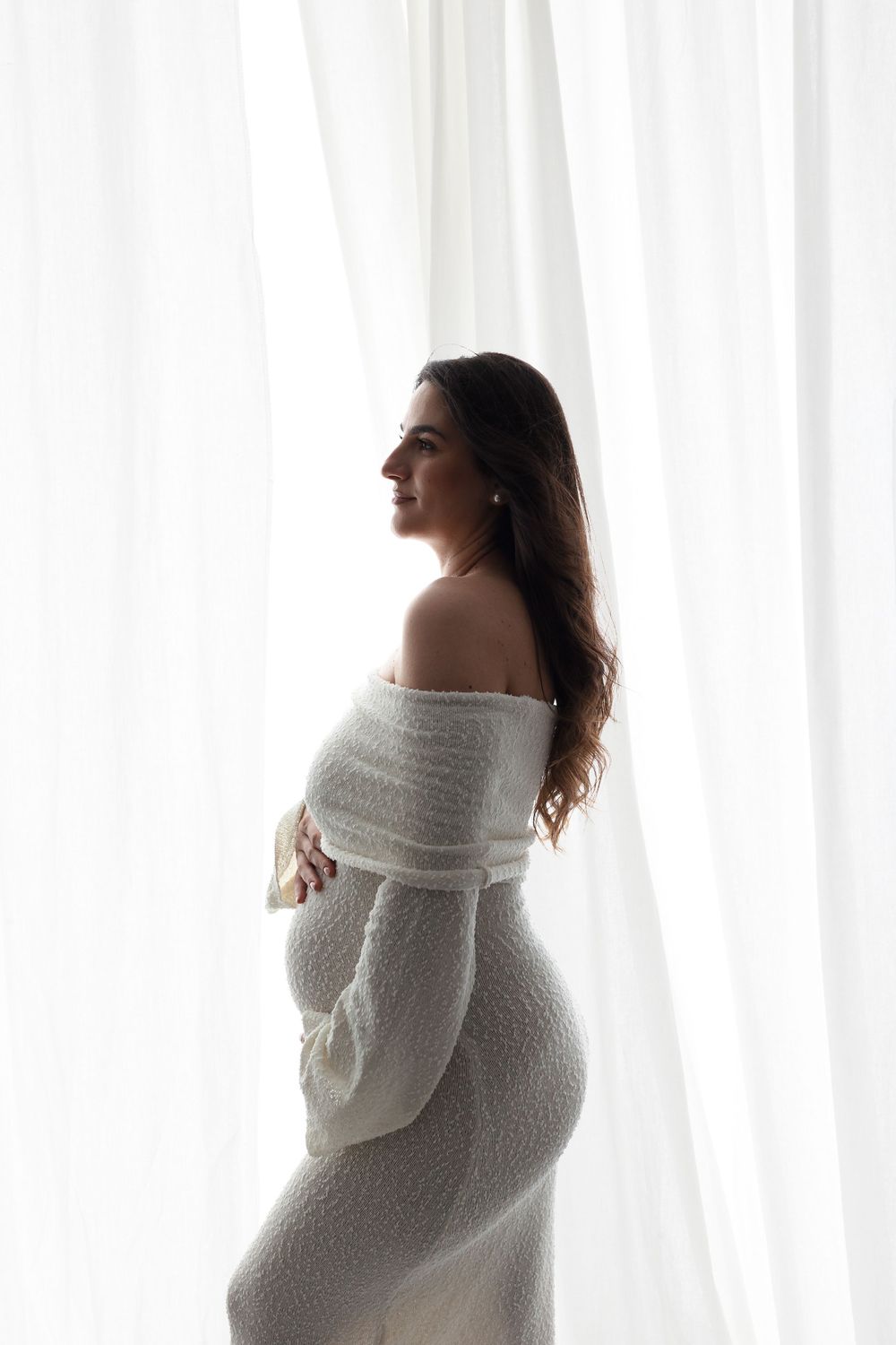 Pregnant woman in white off-shoulder knit dress cradling her baby bump by sheer curtains in soft natural light.