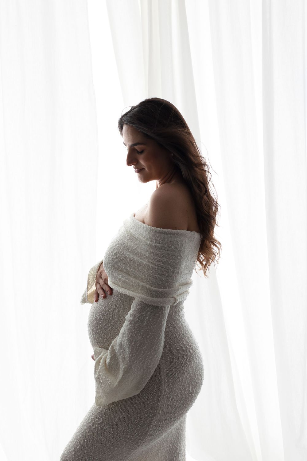 Smiling pregnant woman in white off-shoulder knit dress cradling her baby bump by sheer curtains.