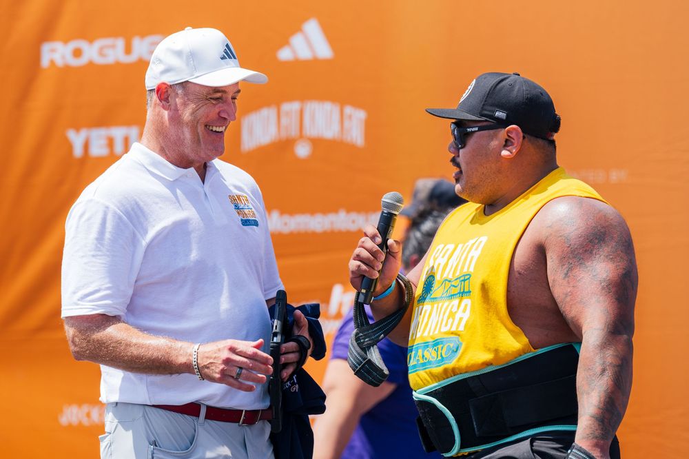 Two people stand in conversation against an orange backdrop at what appears to be a sporting event.