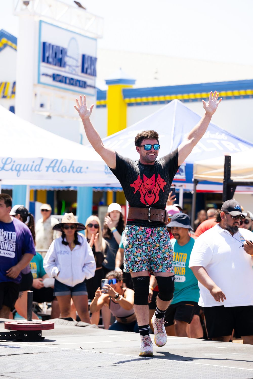 Athlete in black t-shirt and colorful shorts jumps with arms raised during outdoor street competition with crowd watching.
