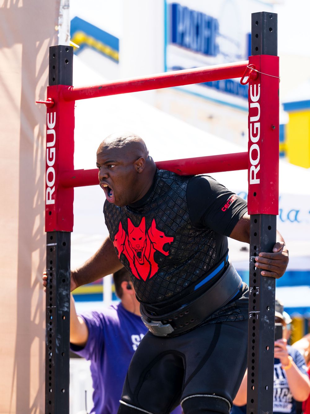 Ajuna Rwakatare in dark clothing performs a heavy lift with red Rogue brand weightlifting equipment during competition.