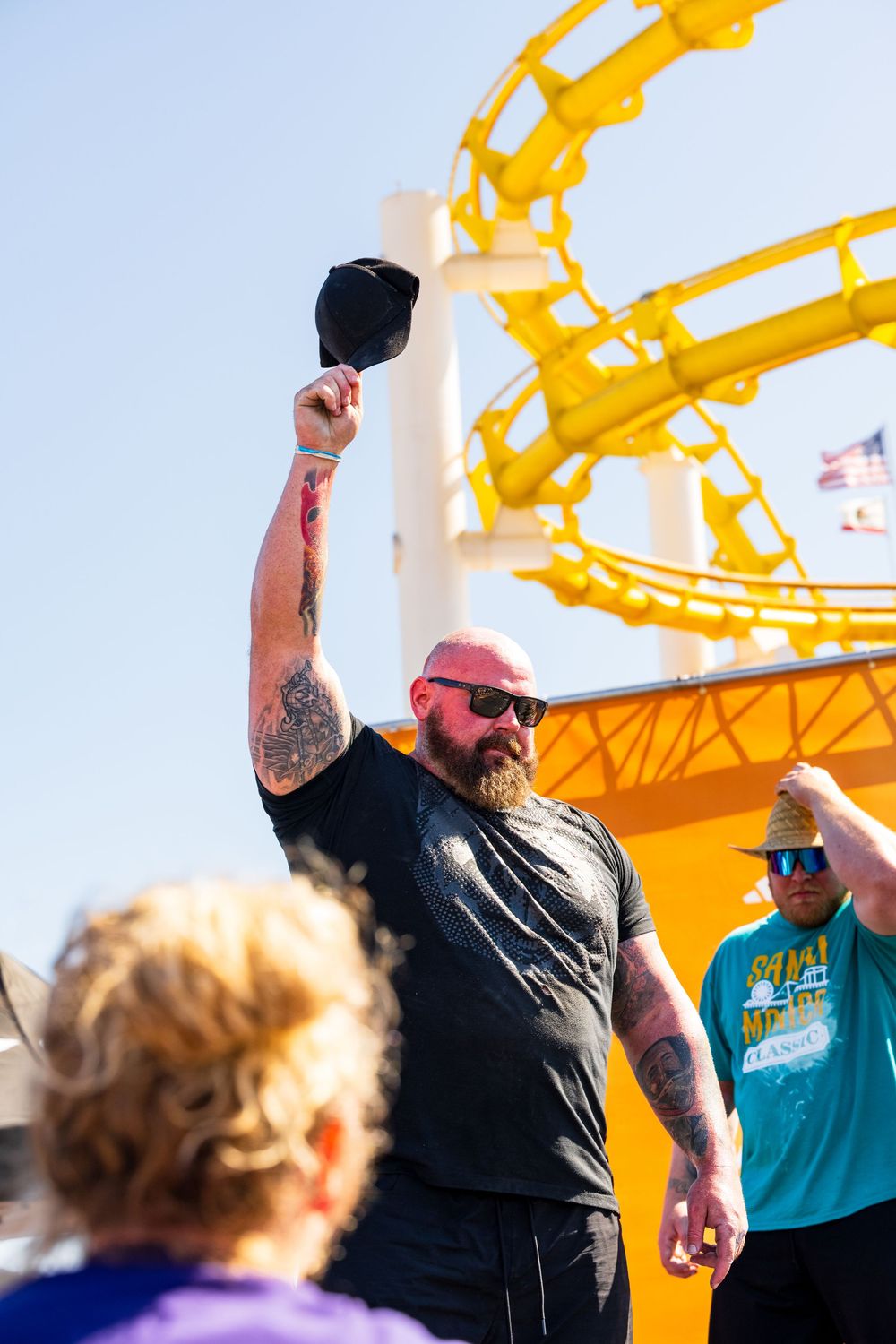 A strongman competitor raises a hat overhead against a bright yellow carnival ride backdrop at Santa Monica Pier.