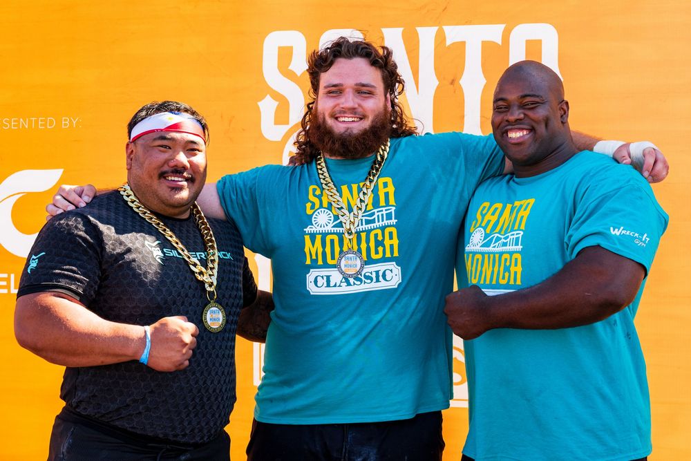 Three people in casual attire posing together and smiling against an orange background with 'SANTO' text Santa Monica's Strongman Event.