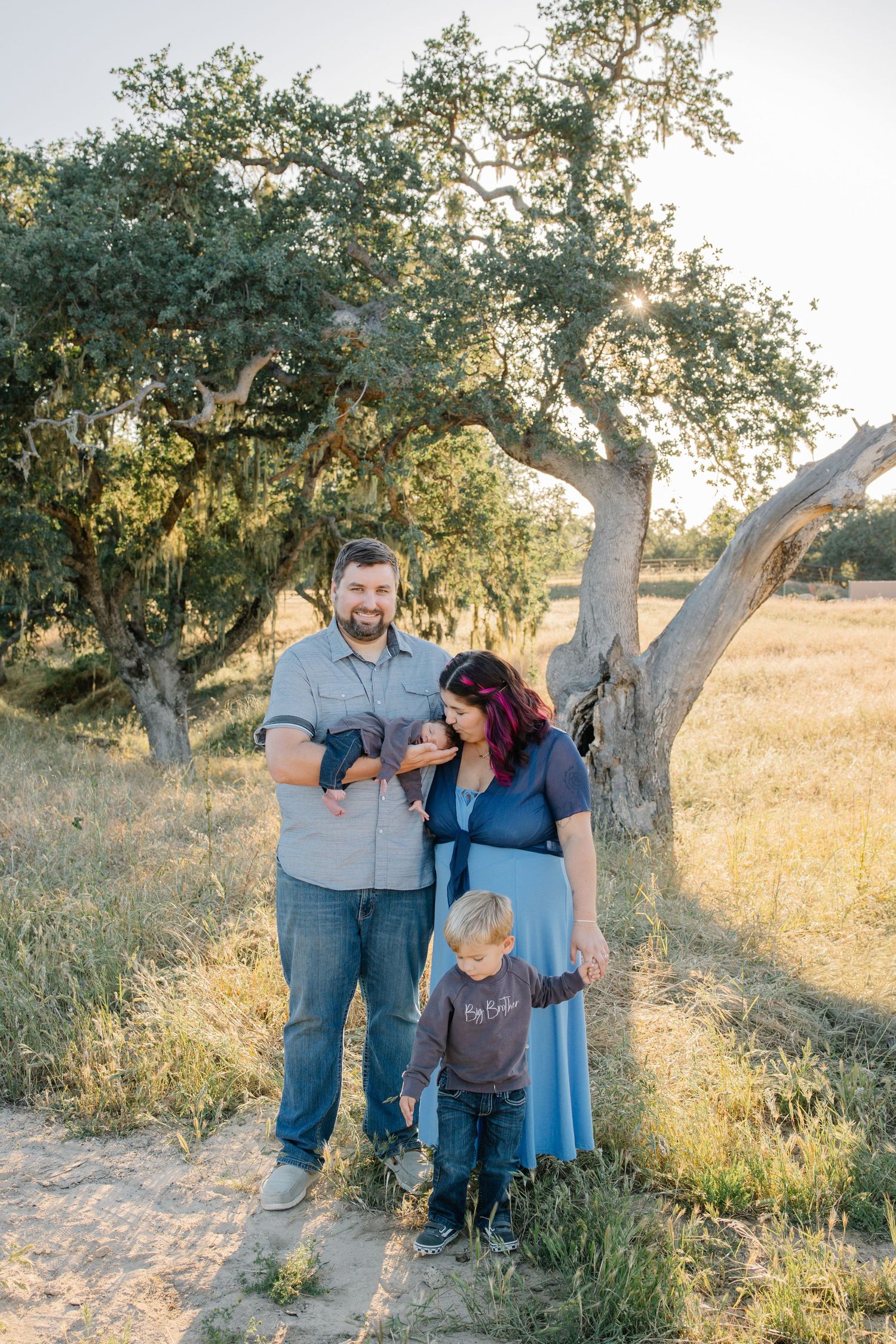 Family of four pose mom kissing newborn baby in dads arms and toddler standing below in grass and oak trees during sunset, golden hour photo