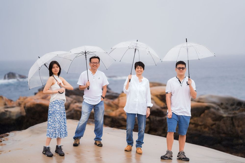 Four people holding white umbrellas pose on a coastal walkway with rocky ocean views on a misty day.
