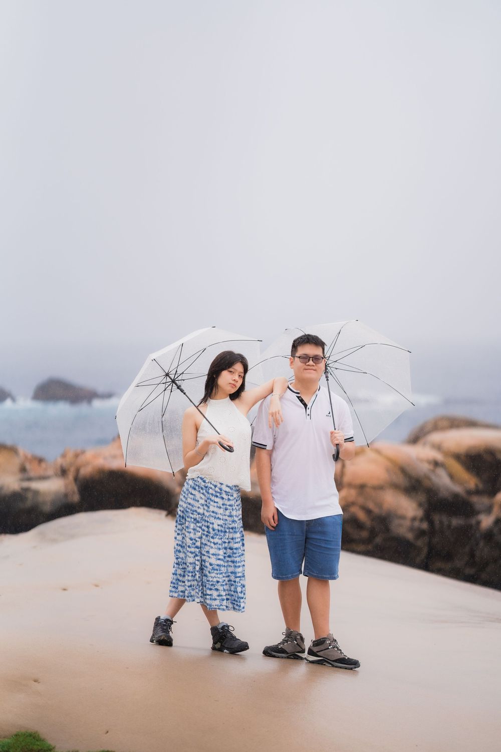 Asian couple sharing white umbrellas while posing together on a sandy coastal path with rocky shoreline.