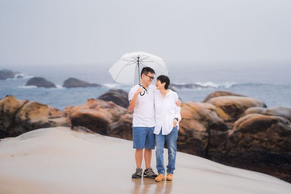 Couple sharing umbrella on rocky beach in misty weather, wearing white shirts and jeans near ocean waves.