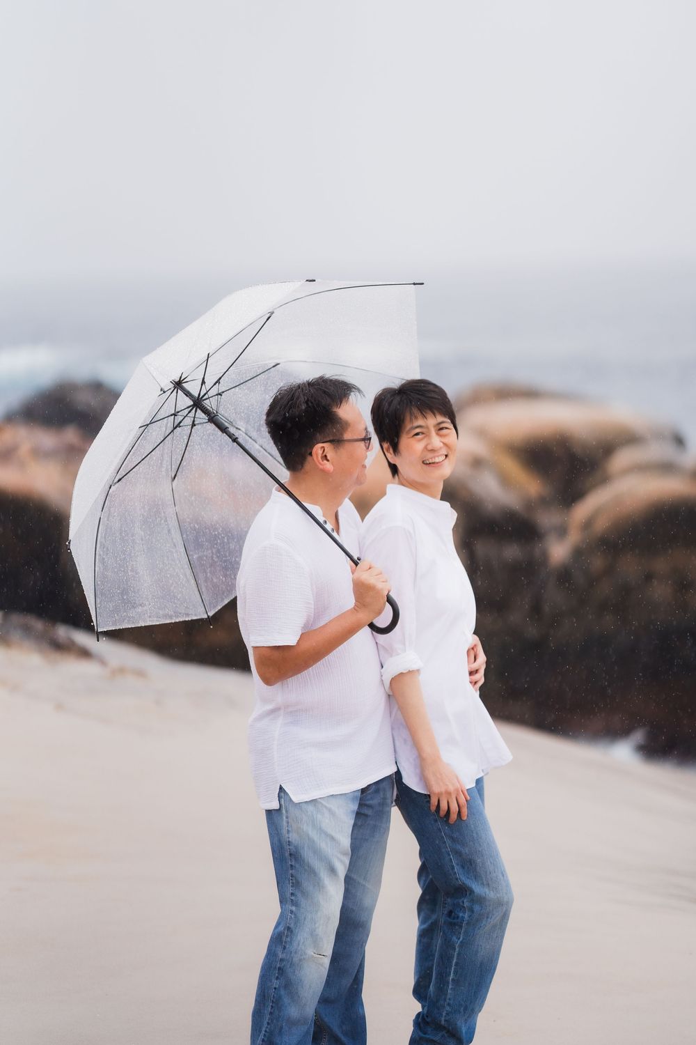 Smiling Asian couple sharing a clear umbrella while walking on a sandy beach with rocky background.