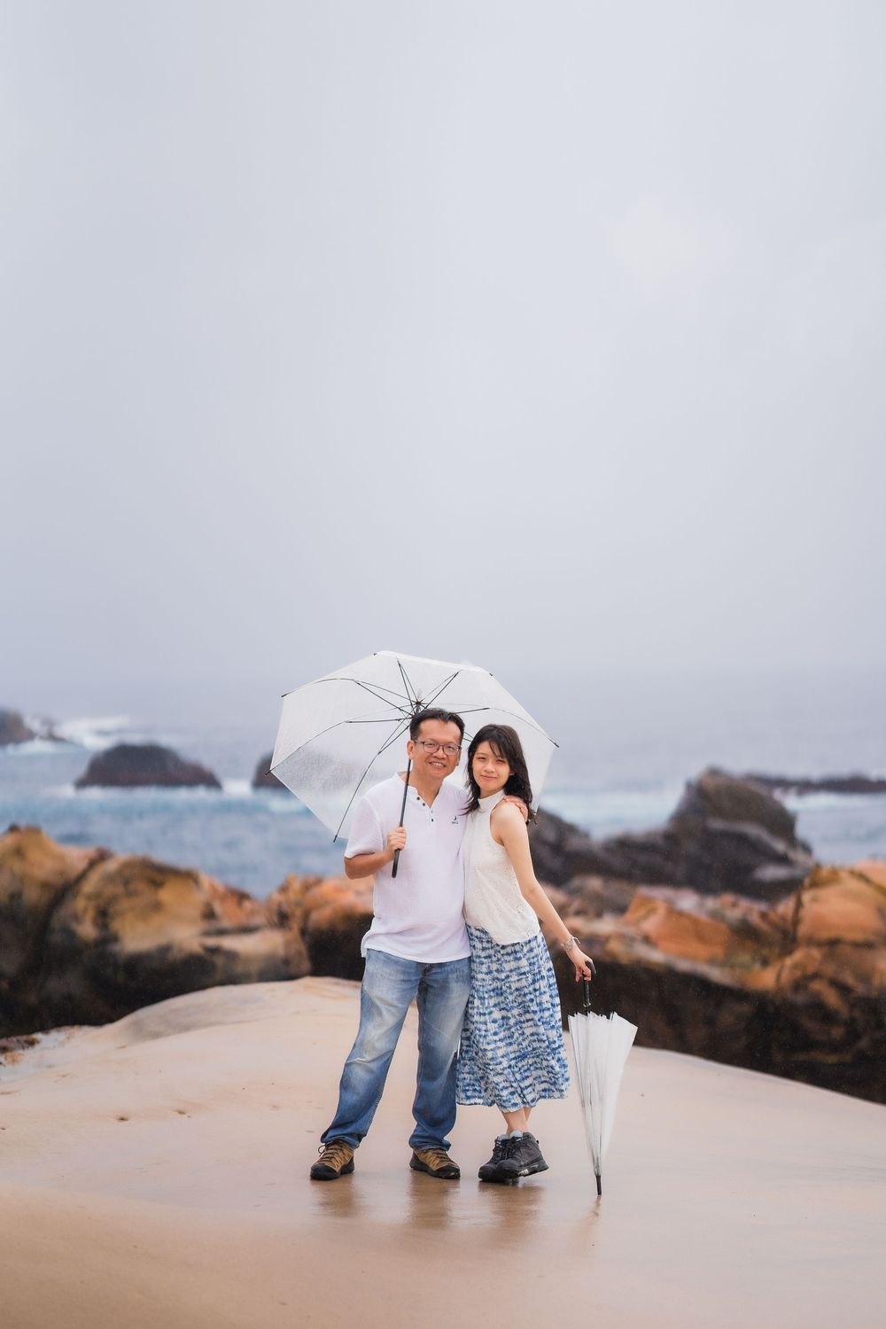 Couple standing on a beach under a white umbrella on a cloudy day, with rocky shores and ocean waves in the background.