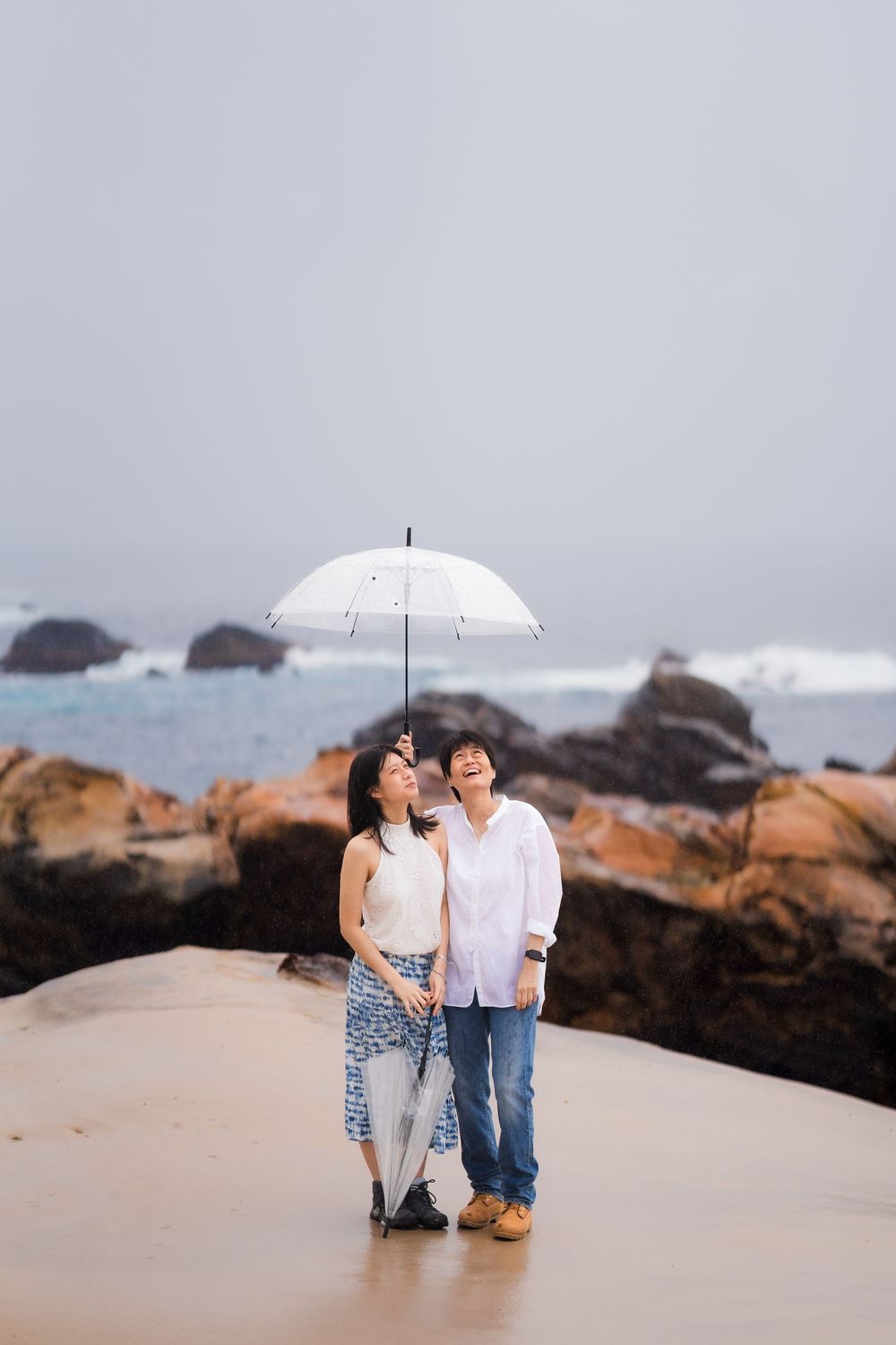 Asian couple standing under a white umbrella on a sandy beach looking up on a cloudy day.