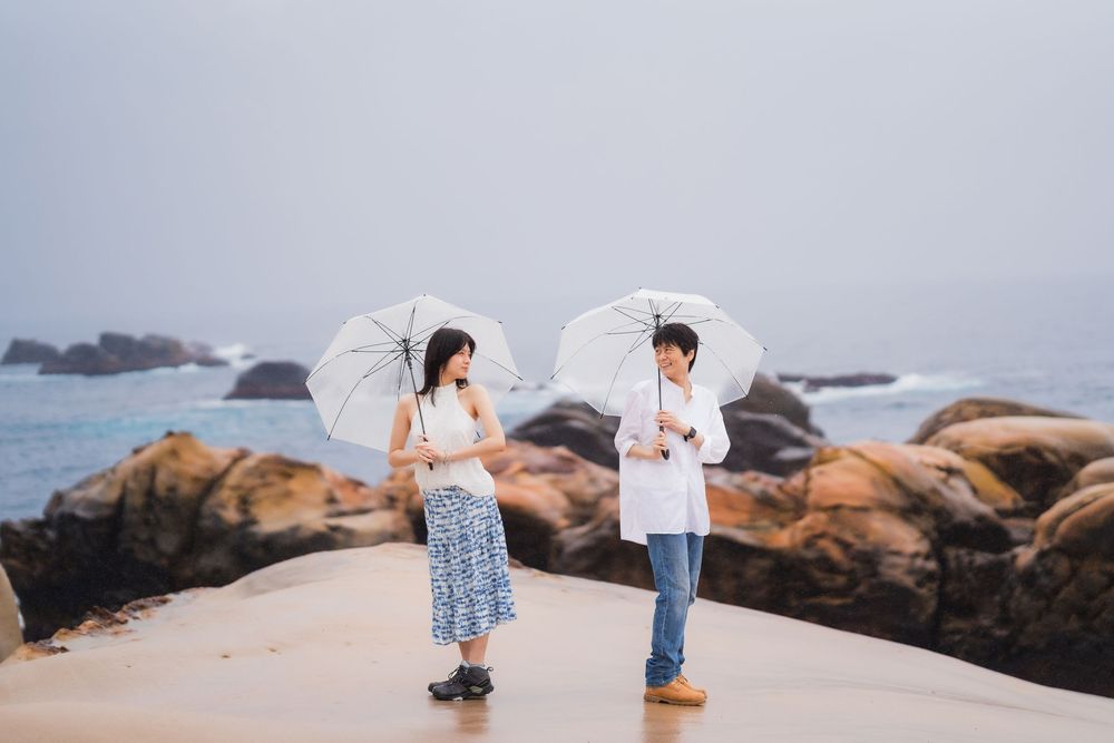 Two people holding white parasol umbrellas stand on a sandy beach with rocky shores and misty ocean in the background.