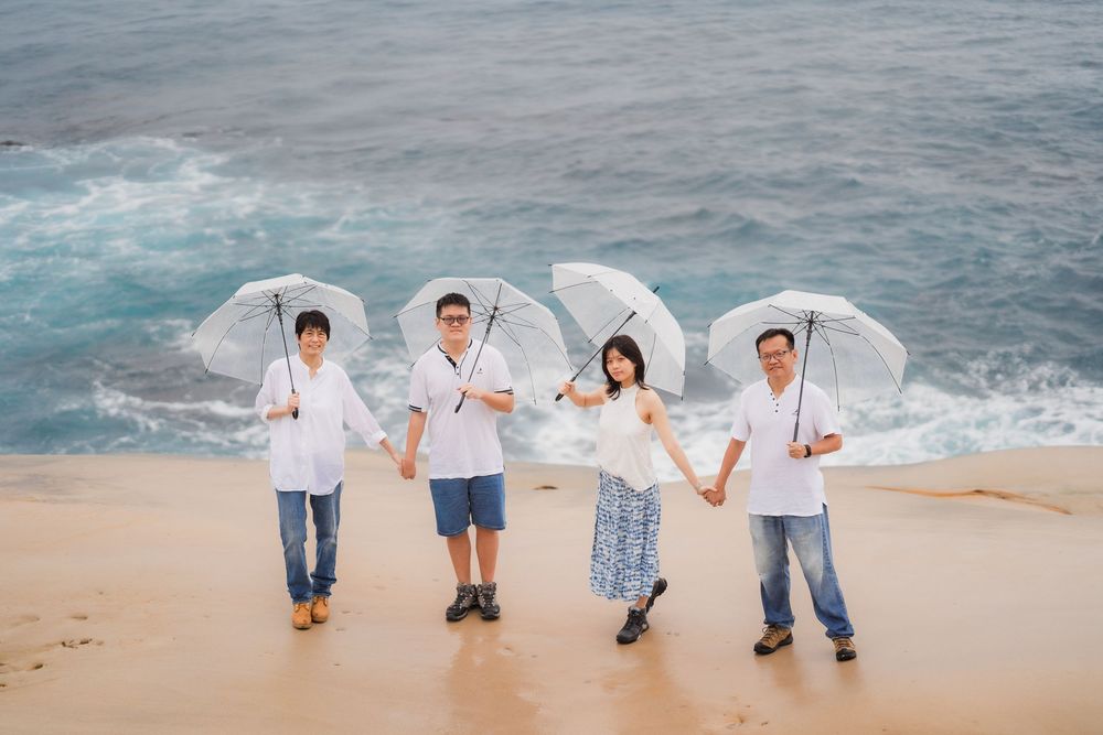 Family of four holding white umbrellas posing on a sandy beach cliff with ocean waves in the background.