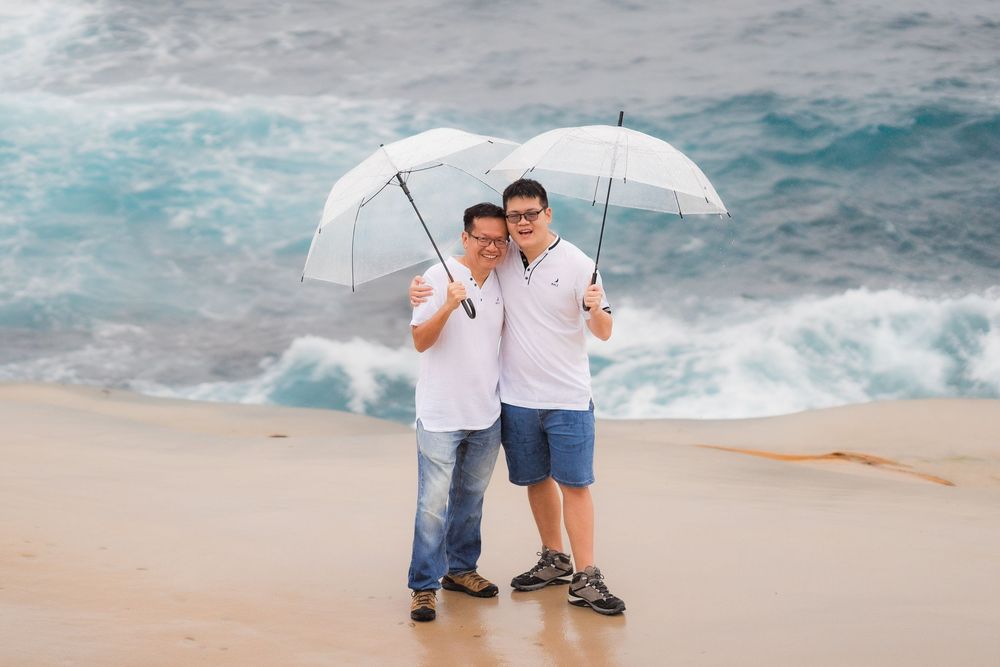 Two men sharing white umbrellas smile together on a sandy beach with dramatic ocean waves behind them.