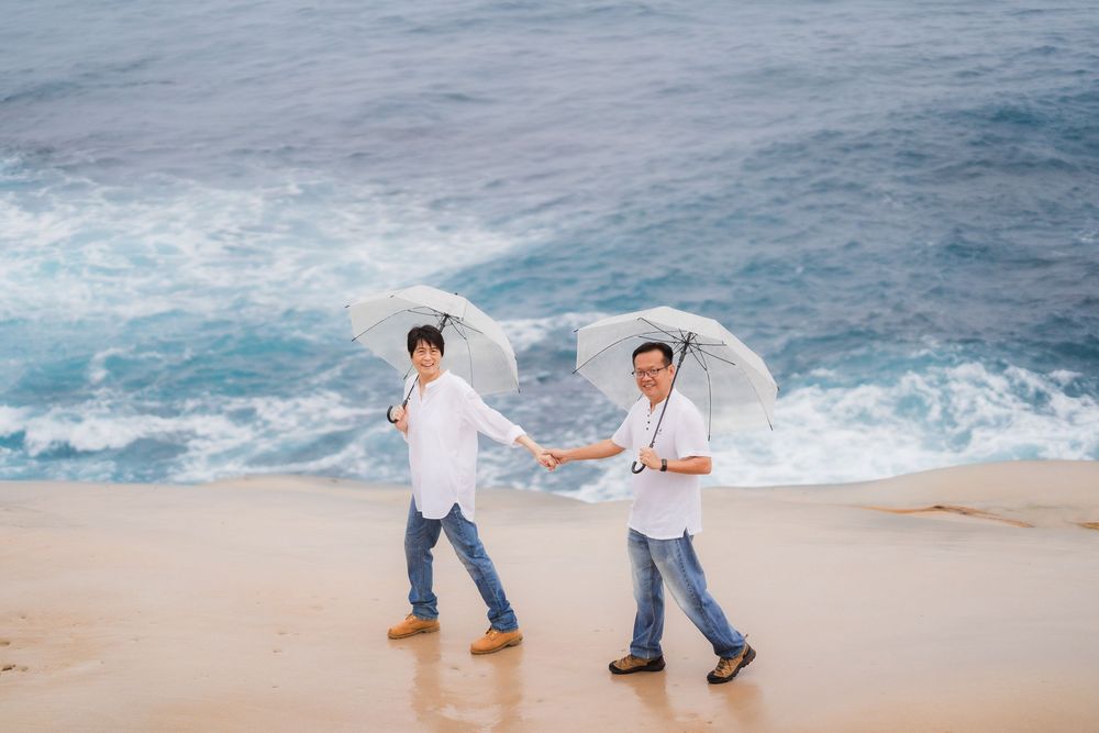 Two men in white shirts holding white umbrellas walk hand in hand on a sandy beach with ocean waves behind them.