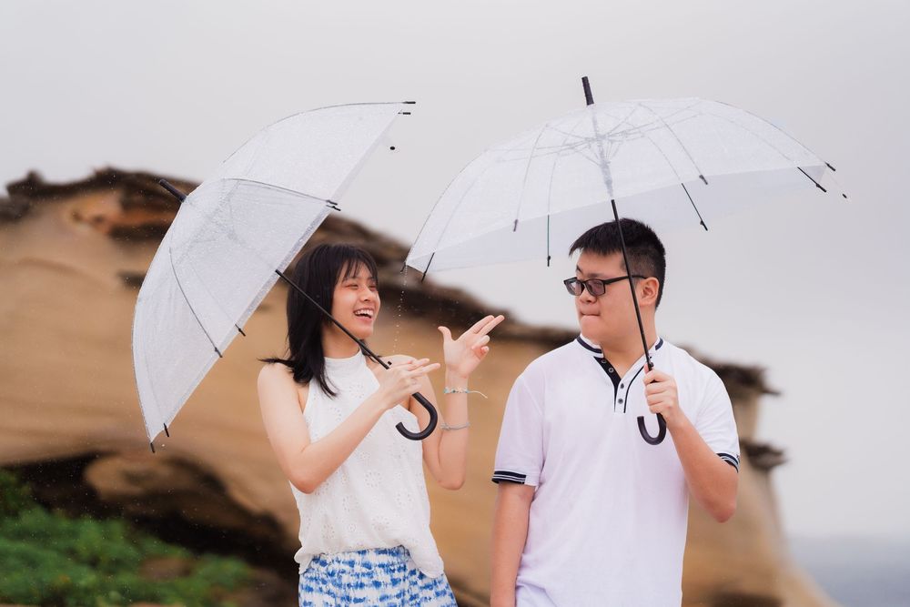 A young Asian couple holding white transparent umbrellas outdoors near rocky formations on a cloudy day.
