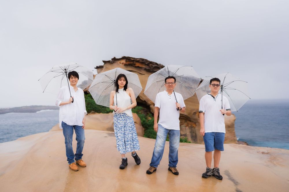 Four people with umbrellas posing on coastal rock formation with ocean view.