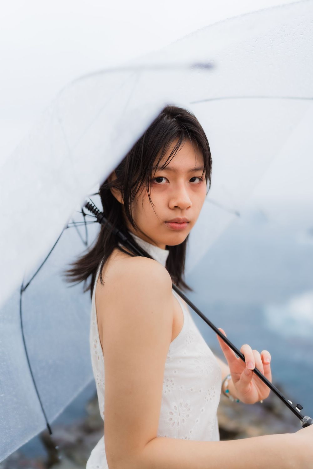 Young Asian woman in white dress holding clear umbrella in rain, looking over shoulder.