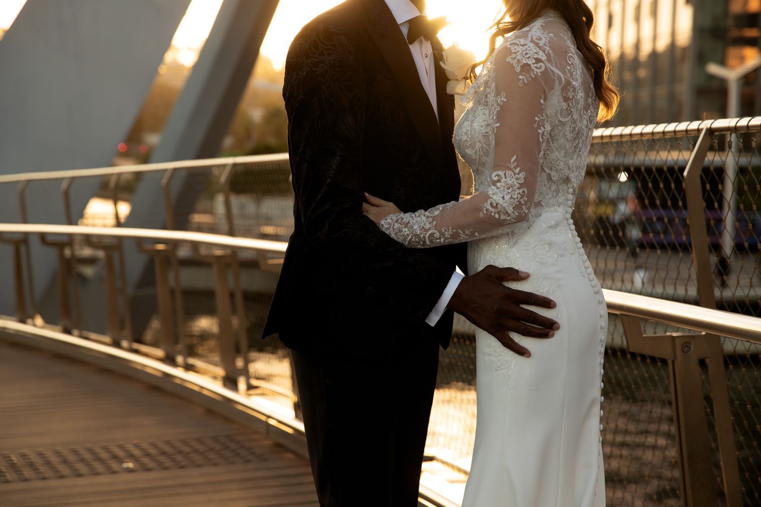 A couple in wedding attire embraces on a bridge at golden hour, bride in lace gown and groom in black tuxedo.