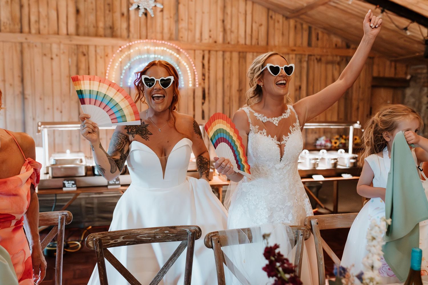Two brides in white gowns celebrate with rainbow fans and heart sunglasses at a rustic barn wedding reception.