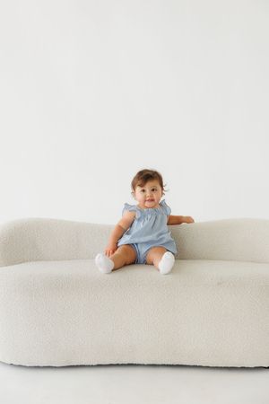 Baby girl sitting on a cream boucle sofa wearing a light blue romper with white socks.